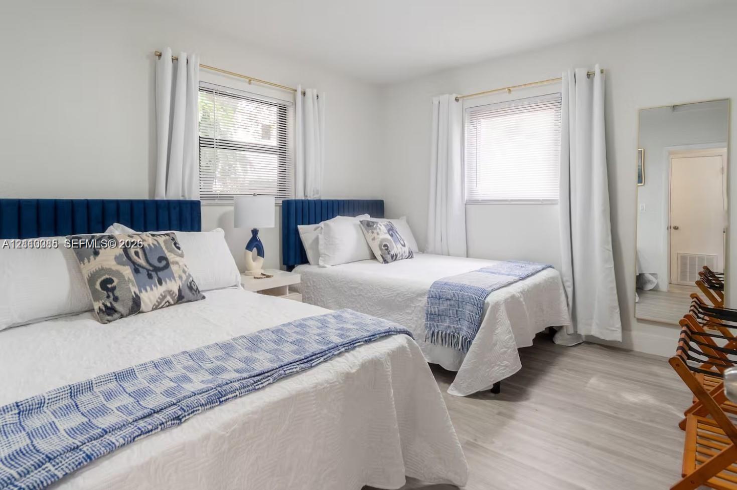Neutral-toned interior featuring wood-finish flooring, two blue upholstered headboards, and dual windows with natural light