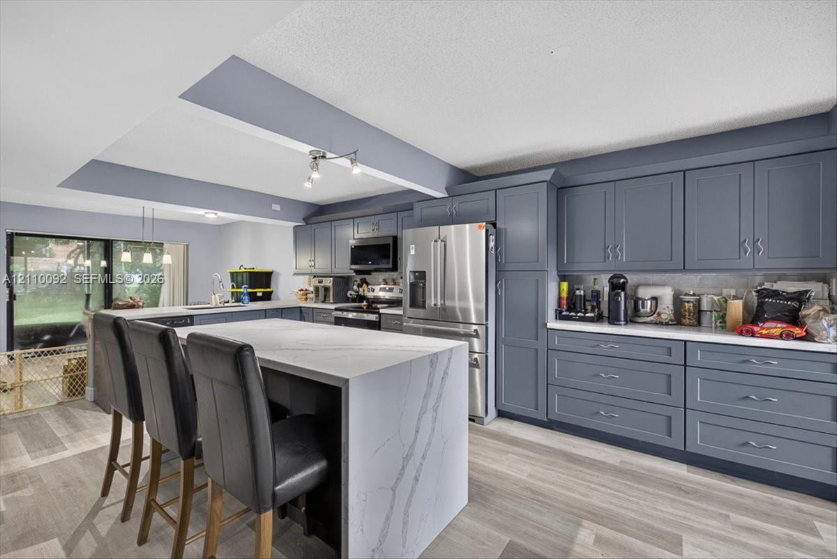 Kitchen featuring shaker-style cabinetry, a waterfall-edge center island with quartz countertops, stainless steel appliances, and wood-finish flooring