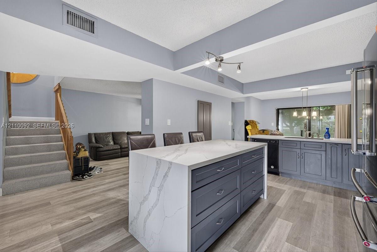 Kitchen featuring an oversized island with a stone-finish waterfall countertop, dark cabinetry, wood-finish flooring, and multi-bulb pendant lighting