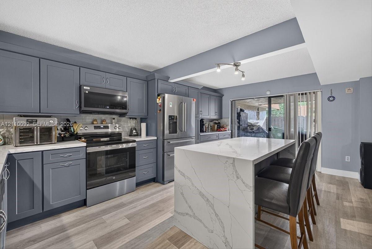 Kitchen featuring shaker-style cabinetry, a waterfall-edge stone island, stainless steel appliances, and wood-finish flooring