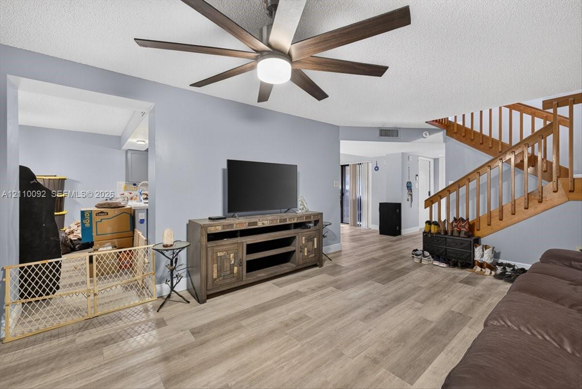 Living area featuring wood-finish flooring, a multi-blade ceiling fan with integrated lighting, and a wooden staircase with balustrades