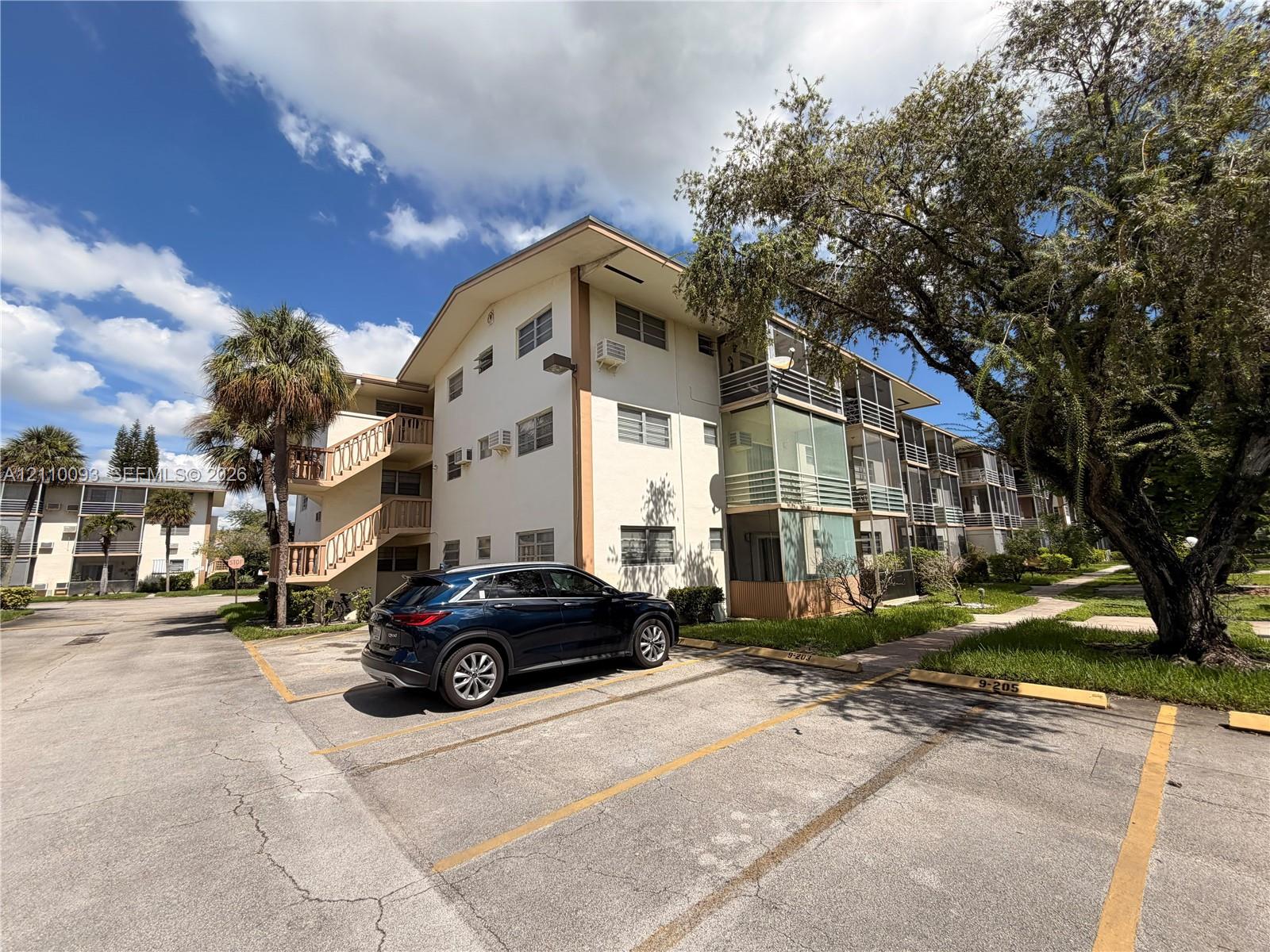 Multi-story exterior featuring a light-colored stucco facade, railed staircases, screened-in balconies, and designated asphalt parking