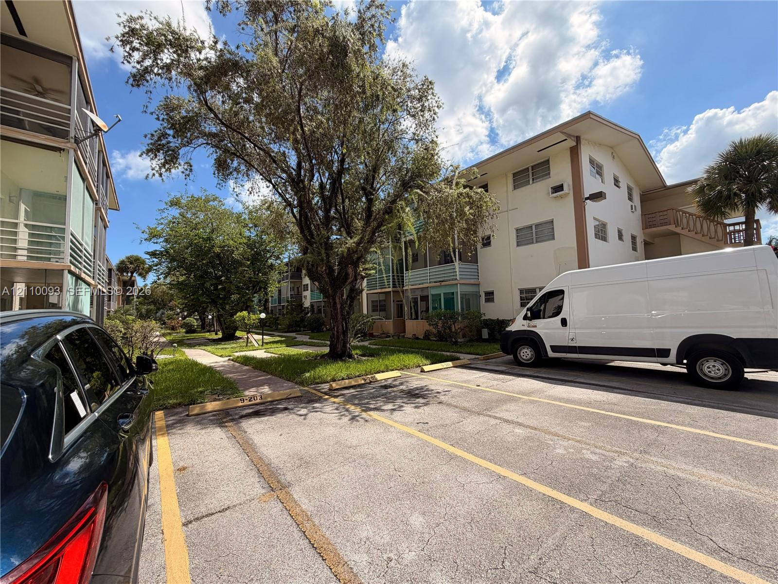 Multi-story exterior featuring a paved parking area with numbered stalls, concrete walkways, screened-in balconies, and mature landscaping Multi-story exterior featuring a paved parking area with numbered stalls, concrete walkways, screened-in balconies, and mature landscaping