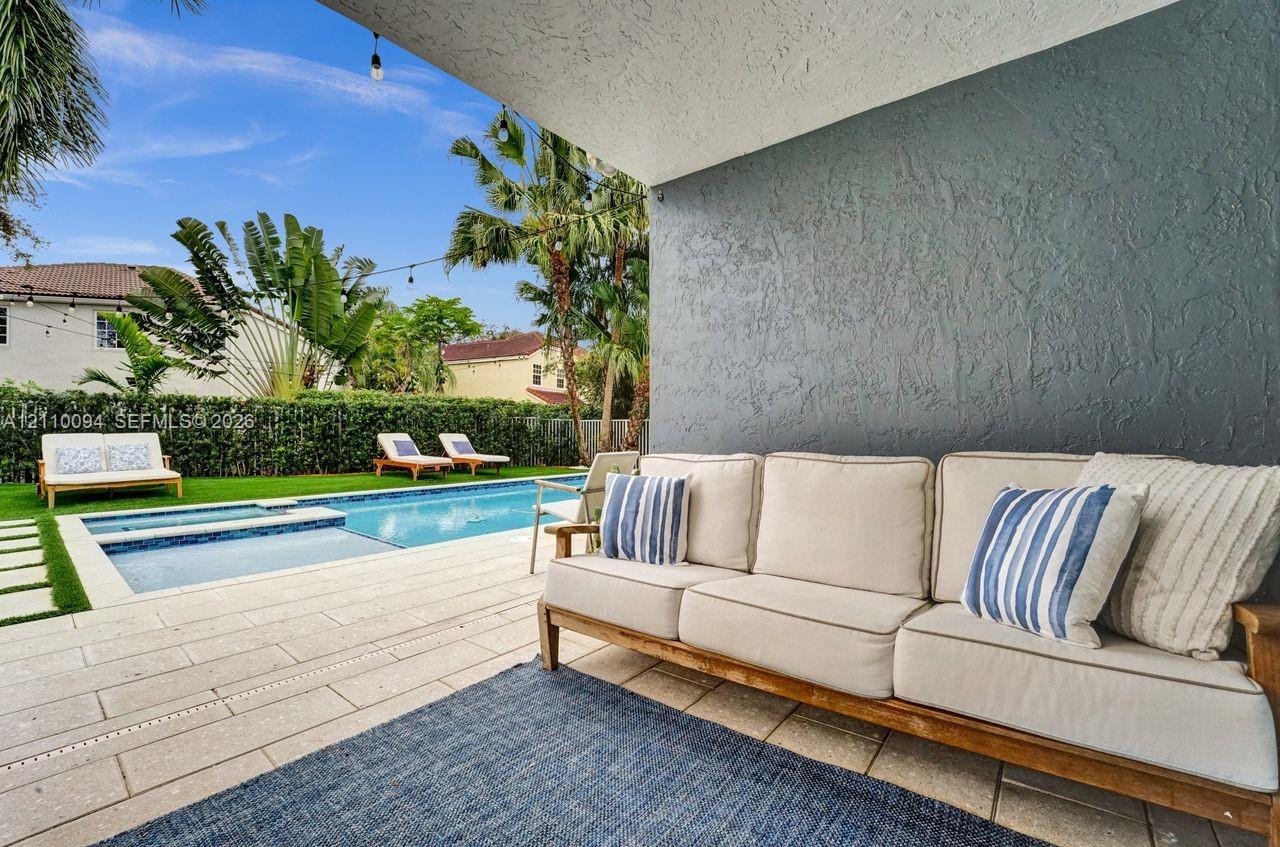 Covered patio featuring a textured grey accent wall, stone-style pavers, and a built-in string lighting system overlooking a rectangular swimming pool with an integrated spa