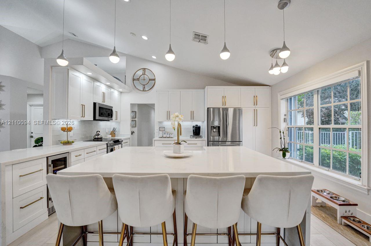 Kitchen featuring white cabinetry with gold-tone hardware, a large center island with a stone-finish countertop, stainless steel appliances, and vaulted ceilings with multiple pendant light fixtures