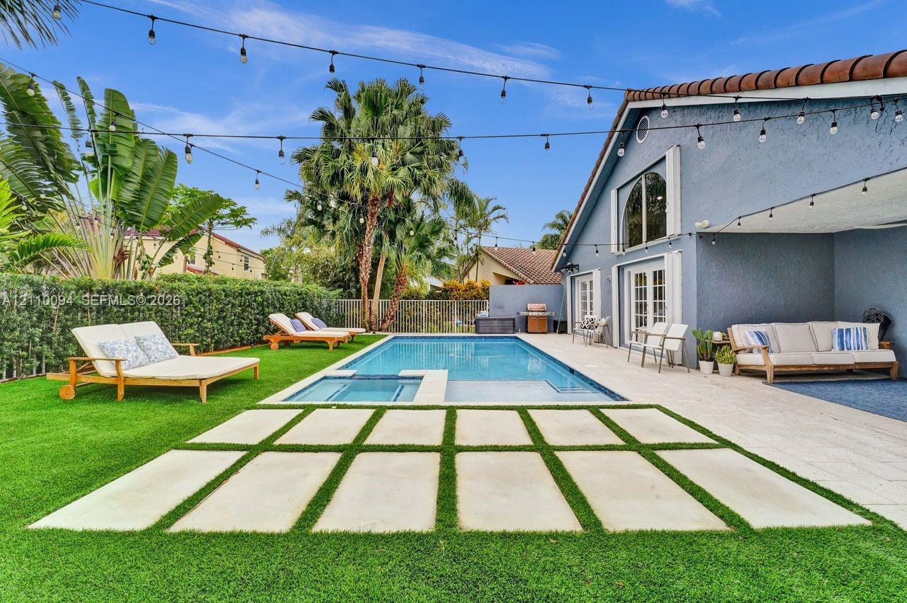 Private backyard featuring an in-ground pool with a built-in spa, paved stone patio with grid inlay, synthetic lawn, and overhead string lighting