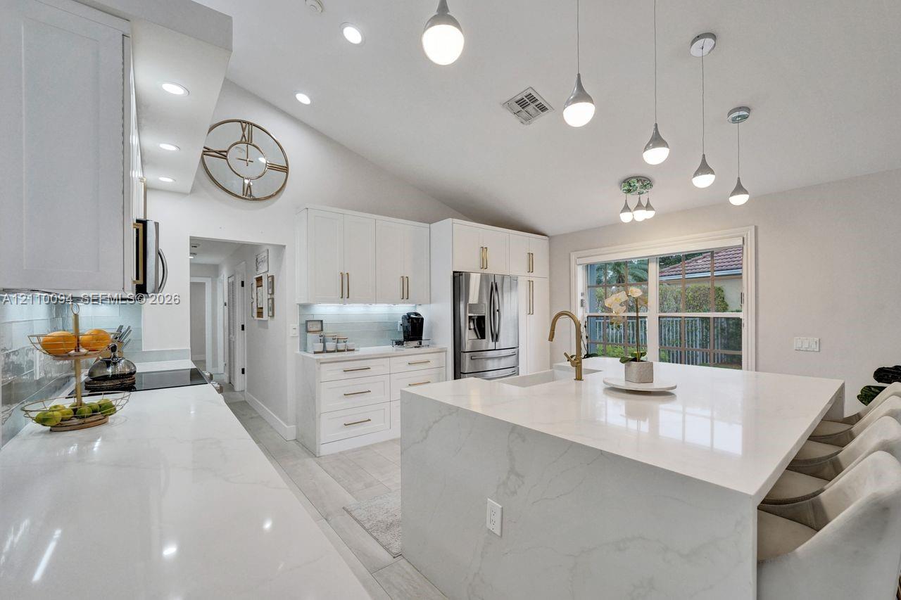 Kitchen featuring white cabinetry with gold-tone hardware, waterfall edge island with stone-finish countertop, under-cabinet lighting, vaulted ceilings, and pendant light fixtures