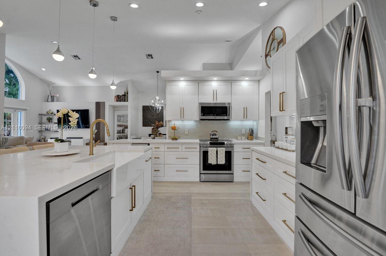 Kitchen featuring white shaker-style cabinetry with gold-toned hardware, quartz countertops, a farmhouse-style apron-front sink, stainless steel appliances, and pendant lighting
