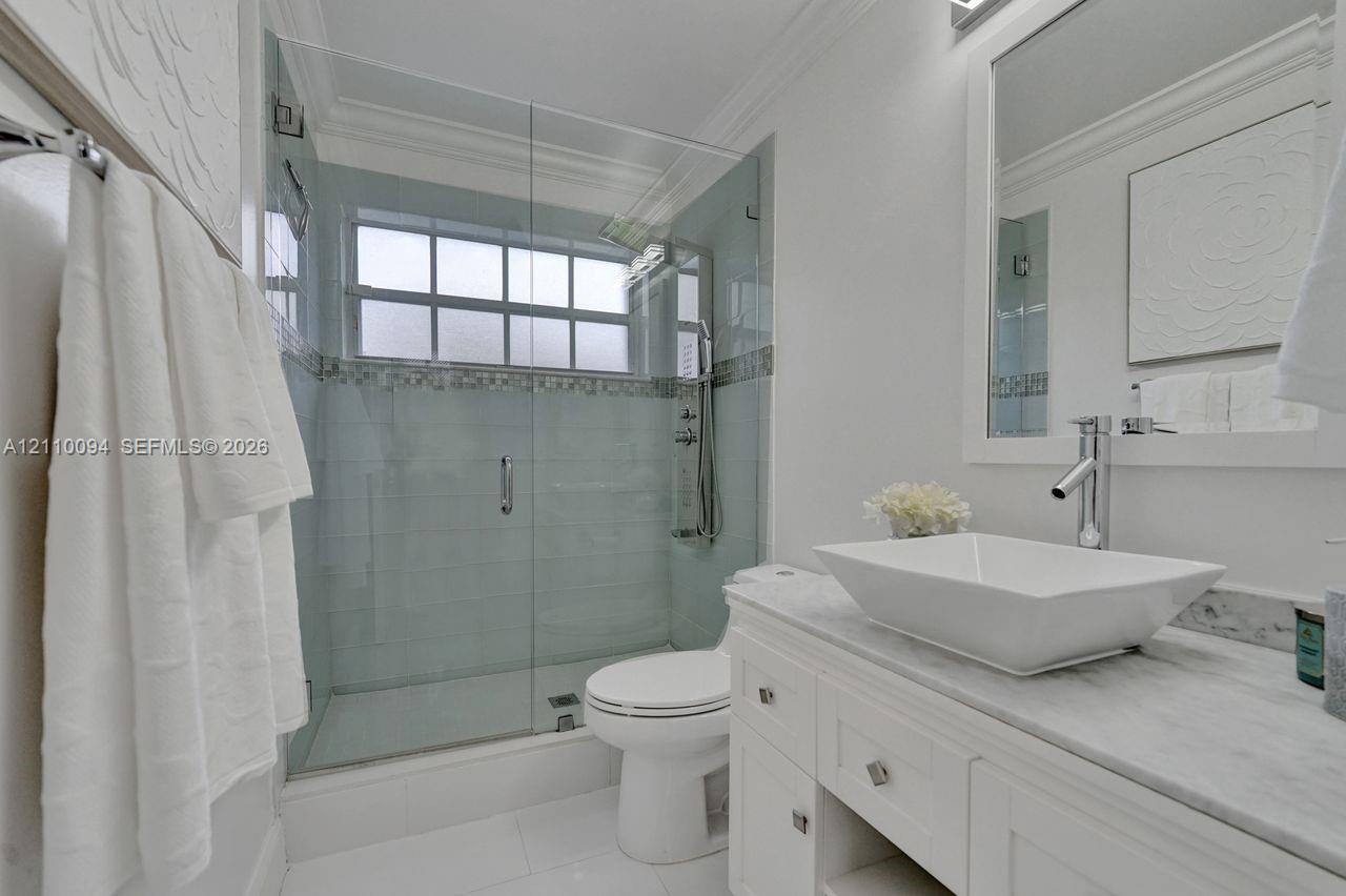 White vanity with a stone countertop, rectangular vessel sink, frameless glass-enclosed shower featuring mosaic tile accents, and a fixed-pane window