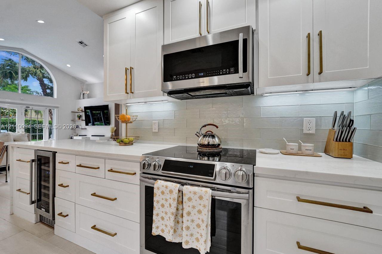 Kitchen featuring white shaker-style cabinetry with brass hardware, solid surface countertops, a subway tile backsplash, stainless steel appliances, and an under-counter wine refrigerator