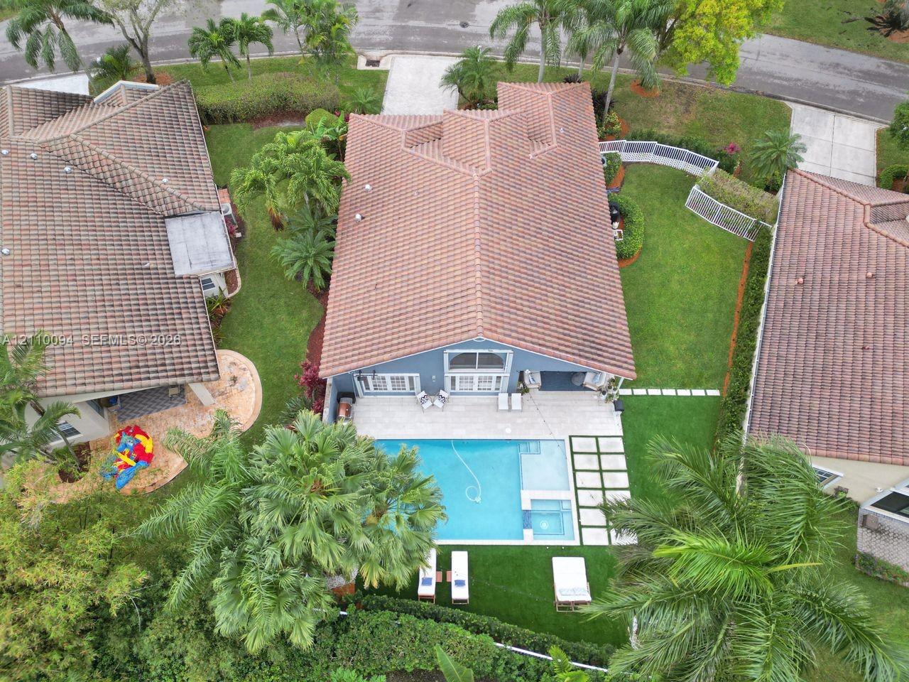 Aerial perspective of a property featuring a tile roof, an in-ground swimming pool with an integrated spa, expansive paver patio, and lush landscaping Aerial perspective of a property featuring a tile roof, an in-ground swimming pool with an integrated spa, expansive paver patio, and lush landscaping