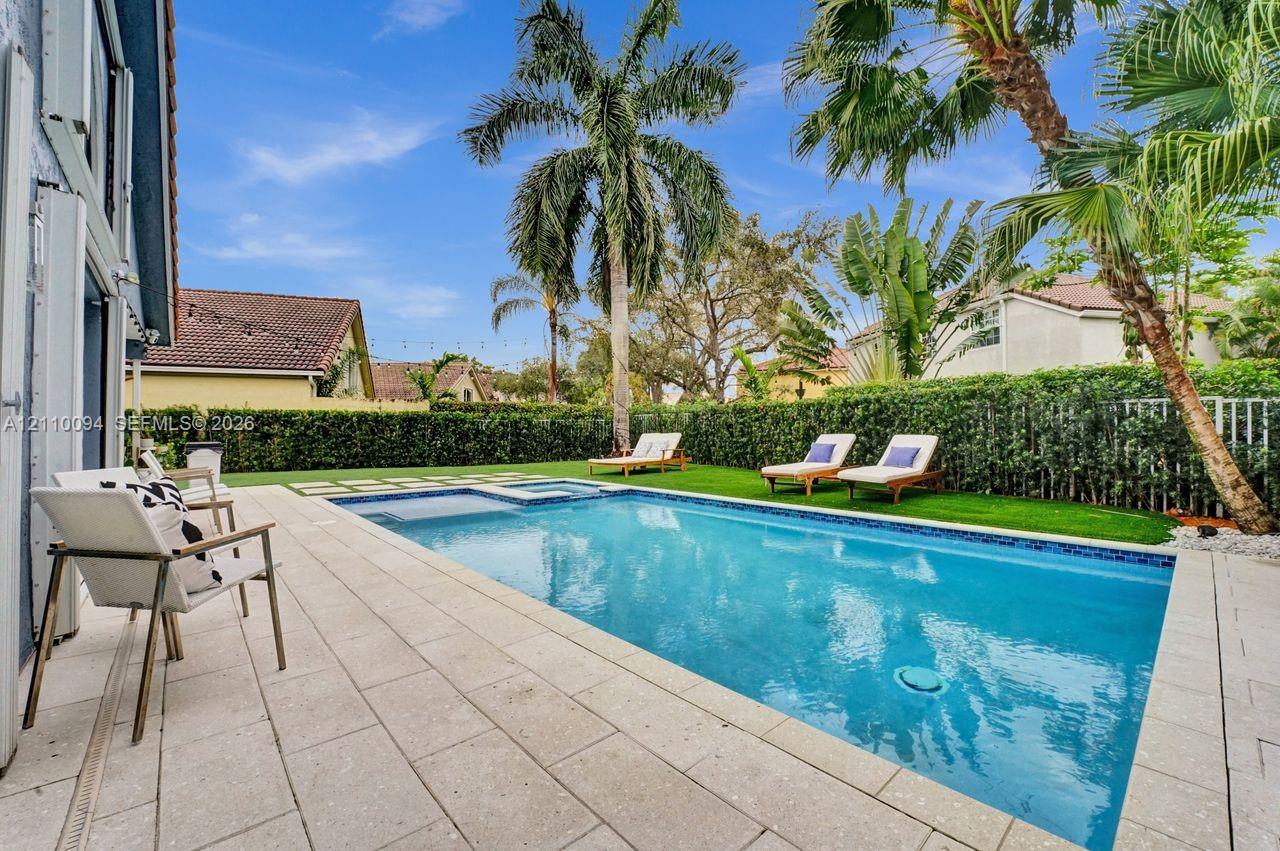 Rectangular swimming pool featuring a built-in spa, stone paver decking, synthetic turf lawn, and mature palm trees