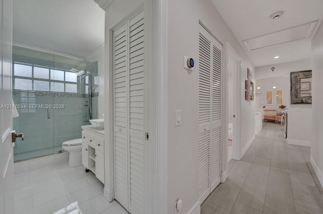 Bathroom featuring a glass-enclosed shower, vessel sink vanity, and white tile flooring, with dual louvered-door closets and wood-finish flooring in the hallway