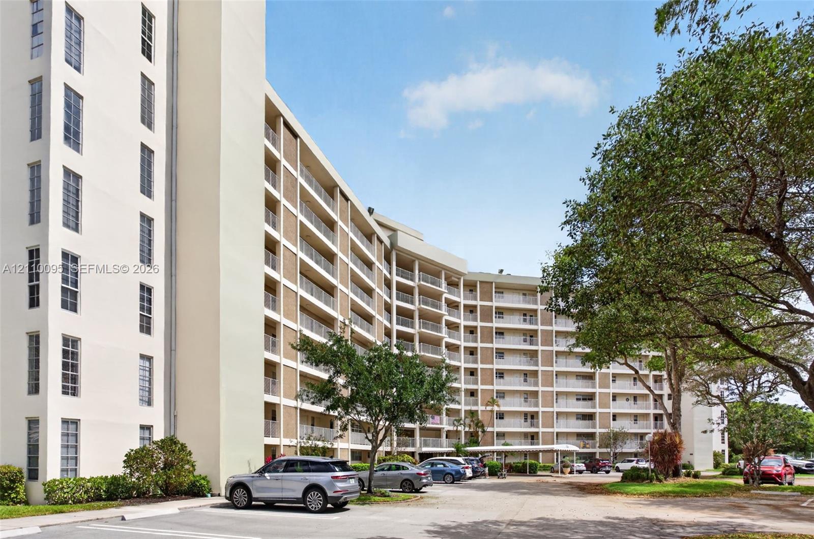 Multi-story residential building exterior featuring stacked balconies, exterior corridor lighting, and a paved parking area with covered parking structures