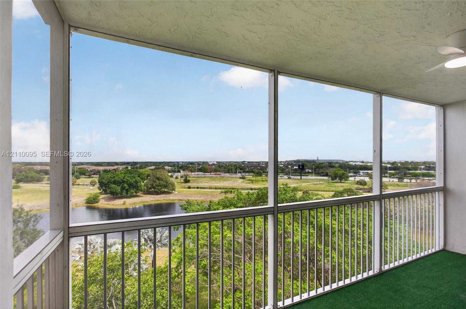 Screened-in balcony featuring white metal railings, a textured ceiling, and an integrated overhead fan, overlooking a pond and landscape