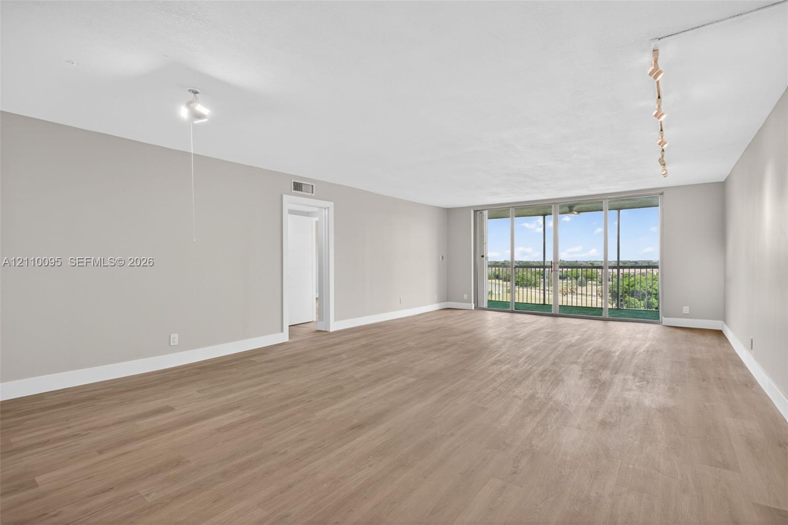 Expansive living area featuring light-toned wood-finish flooring, floor-to-ceiling glass sliding doors, a covered balcony, and adjustable track lighting