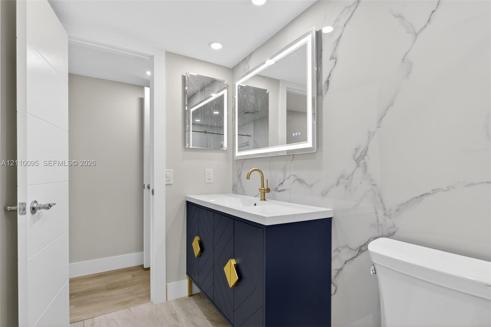 Navy blue vanity with geometric door detailing, brass hardware, integrated sink, large-format stone-look wall cladding, and a rectangular LED-backlit mirror