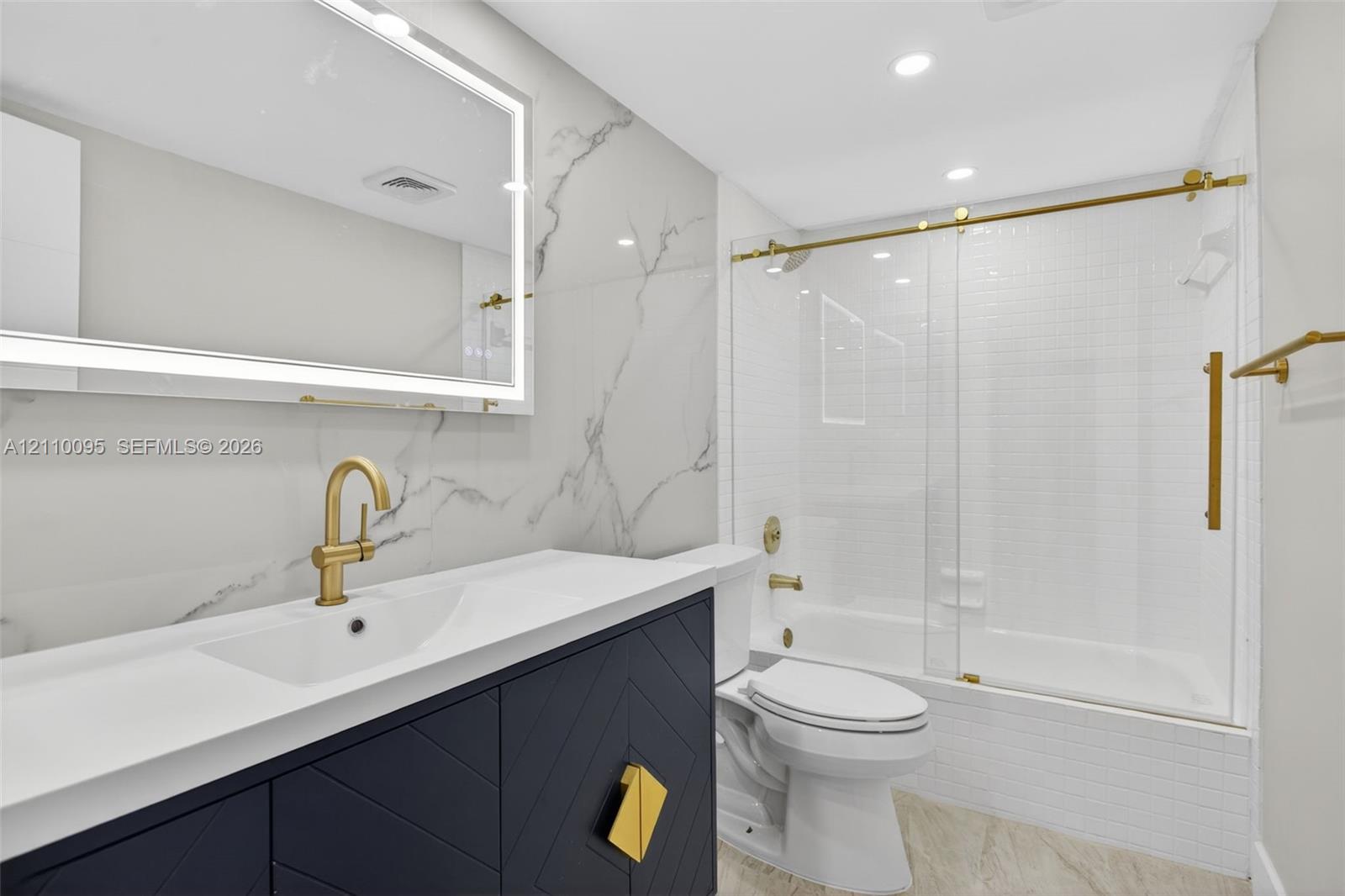Custom navy cabinetry with geometric detailing and gold hardware, white countertop with integrated sink, gold-finished plumbing fixtures, frameless glass shower door with gold trim, and large-format wall tiling with a veined pattern