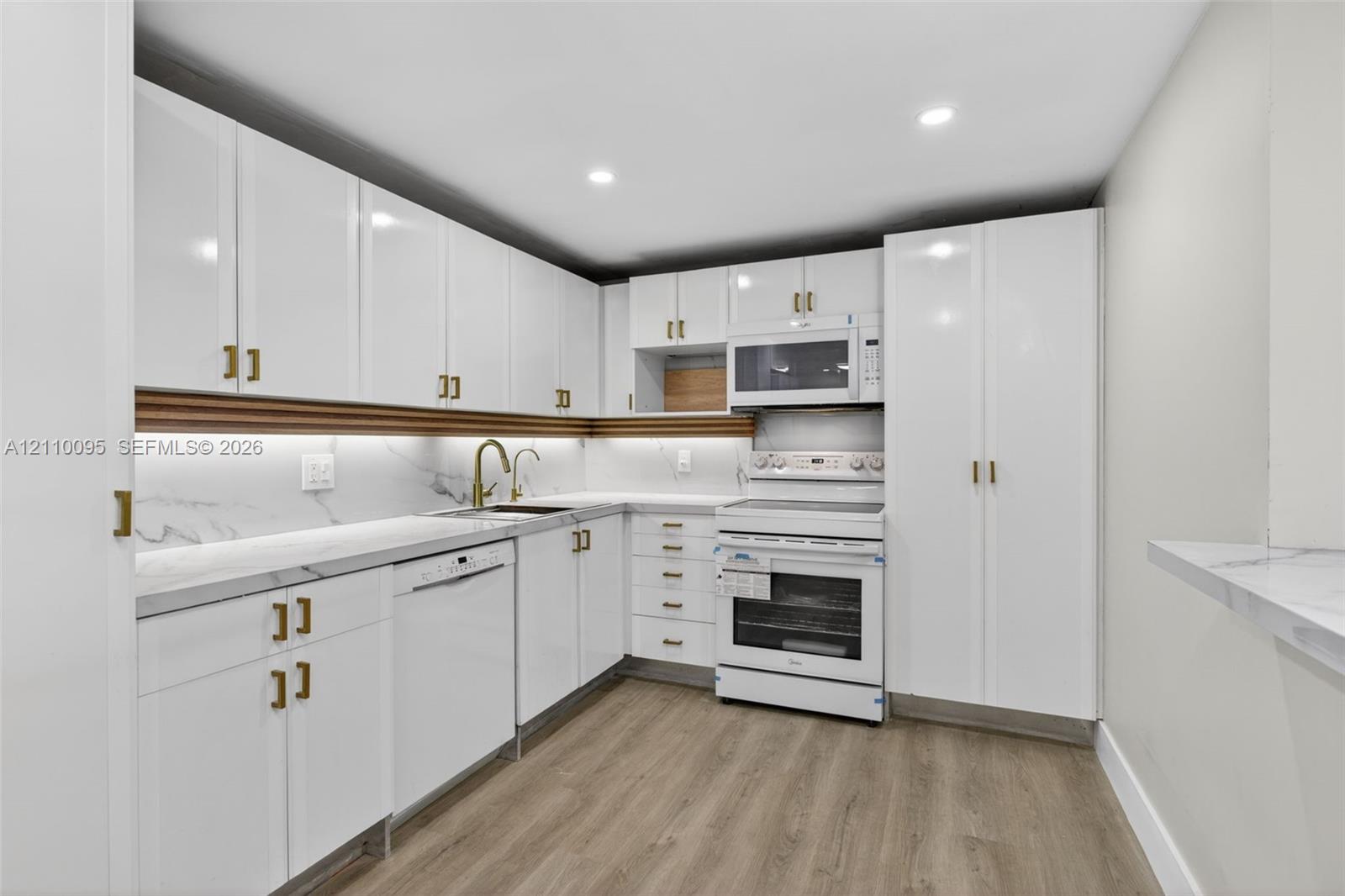 Kitchen featuring white flat-panel cabinetry with gold-tone hardware, stone-surface countertops, integrated under-cabinet lighting, wood-finish flooring, and a built-in microwave