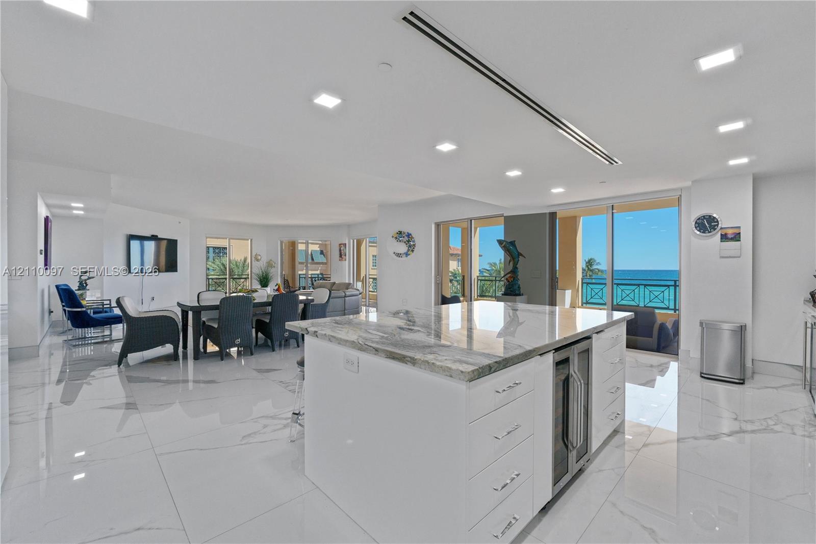 Open-concept kitchen featuring an expansive stone-topped island with an under-counter beverage chiller, high-gloss large-format tile flooring, recessed lighting, and expansive floor-to-ceiling glass doors opening to an exterior balcony