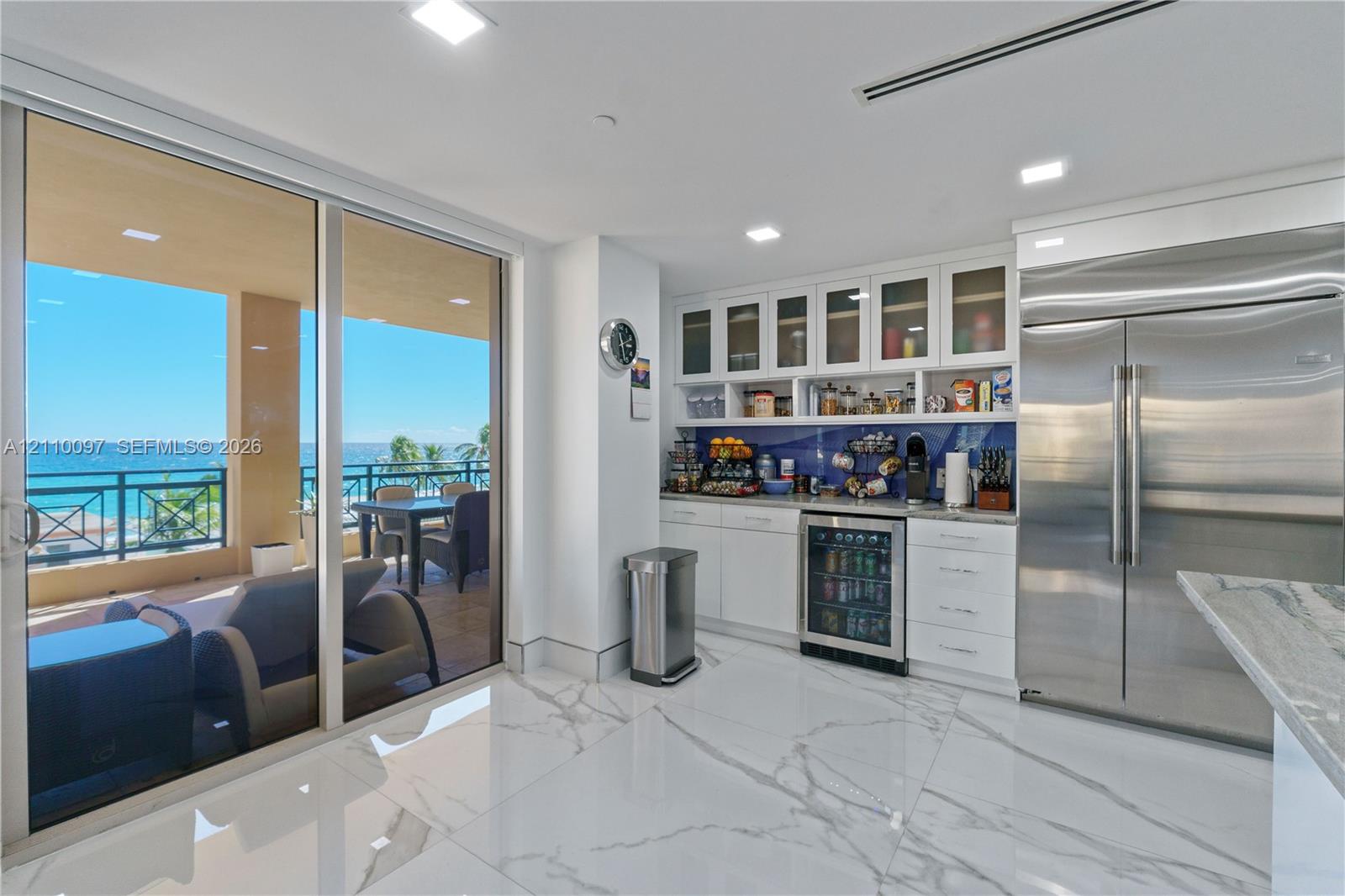 Large-format tile flooring, built-in white cabinetry with glass-front uppers, stainless steel side-by-side refrigerator, under-counter beverage cooler, and expansive floor-to-ceiling sliding glass doors opening to a covered balcony