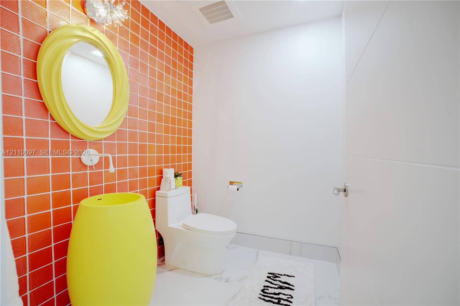 Powder room featuring a floor-to-ceiling orange tile accent wall, a freestanding cylindrical basin, a circular wall-mounted mirror, and stone-finish flooring