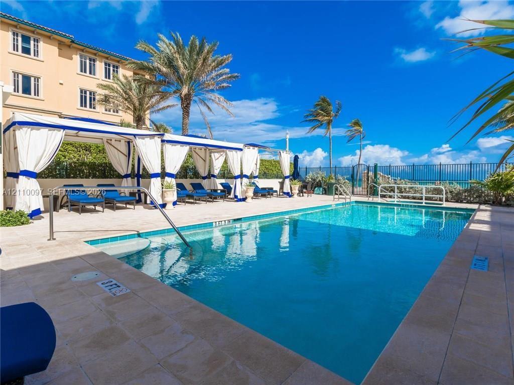 Swimming pool with stone-tiled decking, built-in structural cabanas, metal perimeter fencing, and mature palm trees