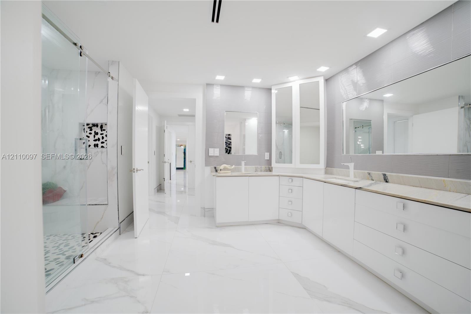Large-format stone tile flooring, frameless glass shower enclosure, custom white cabinetry, stone-topped dual vanity, and recessed ceiling lighting