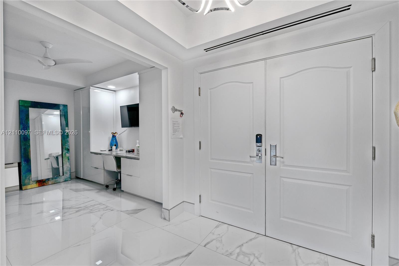 Double entry doors with arch-top panels, large-format tile flooring with marble-finish veining, recessed tray ceiling, and built-in cabinetry featuring integrated workspace Double entry doors with arch-top panels, large-format tile flooring with marble-finish veining, recessed tray ceiling, and built-in cabinetry featuring integrated workspace