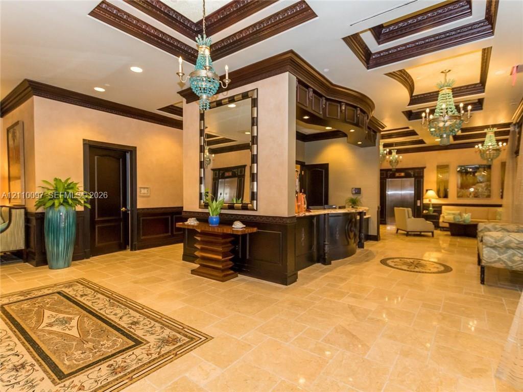 Grand lobby featuring coffered ceilings with dark wood molding, natural stone flooring with inlaid medallion accents, custom built-in reception desk, decorative crystal chandeliers, and wall-mounted mirrors