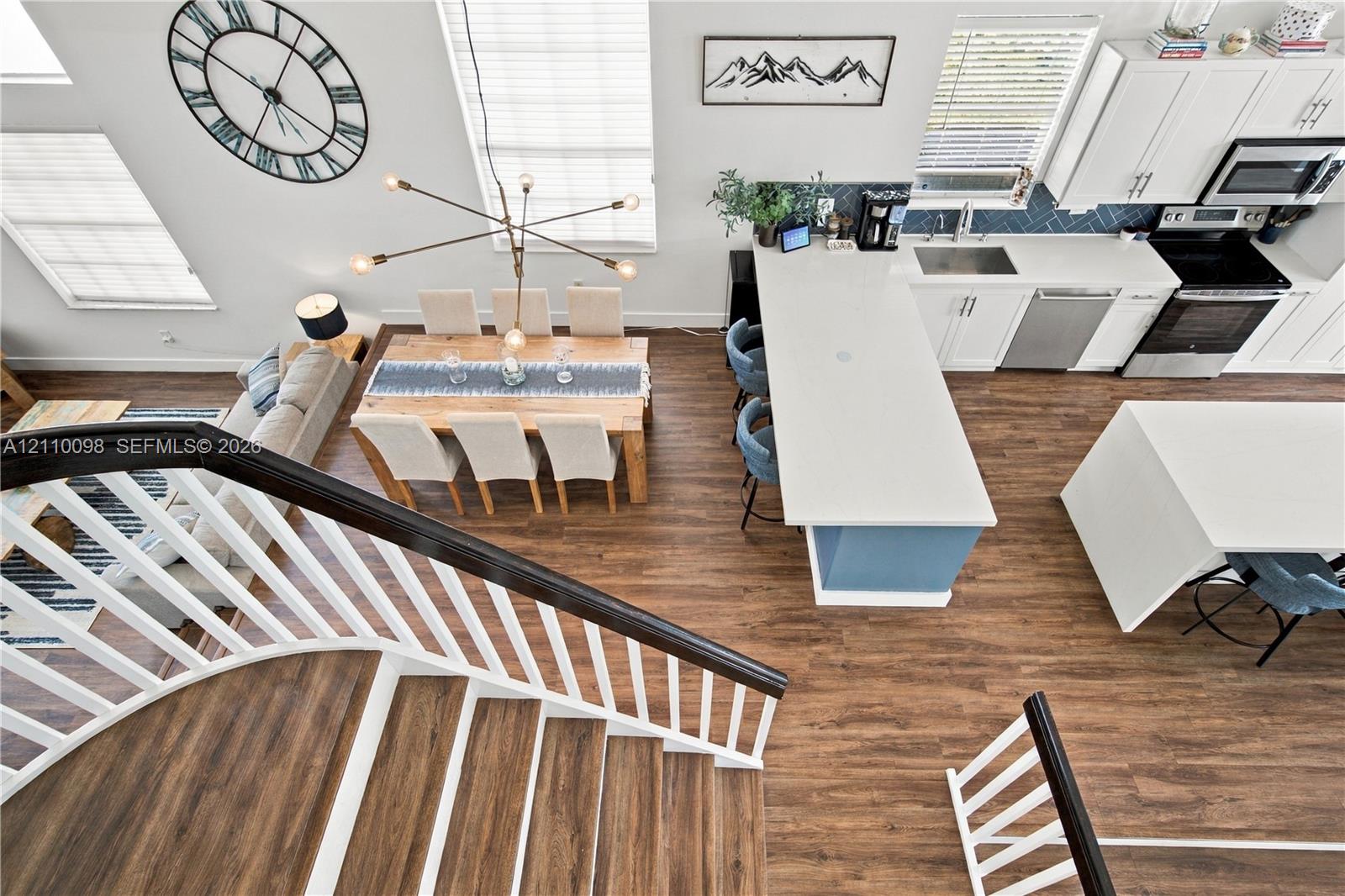 Open floor plan featuring wood-finish flooring, a kitchen with a quartz-topped island, white cabinetry, herringbone backsplash, and a staircase with a dark-stained handrail