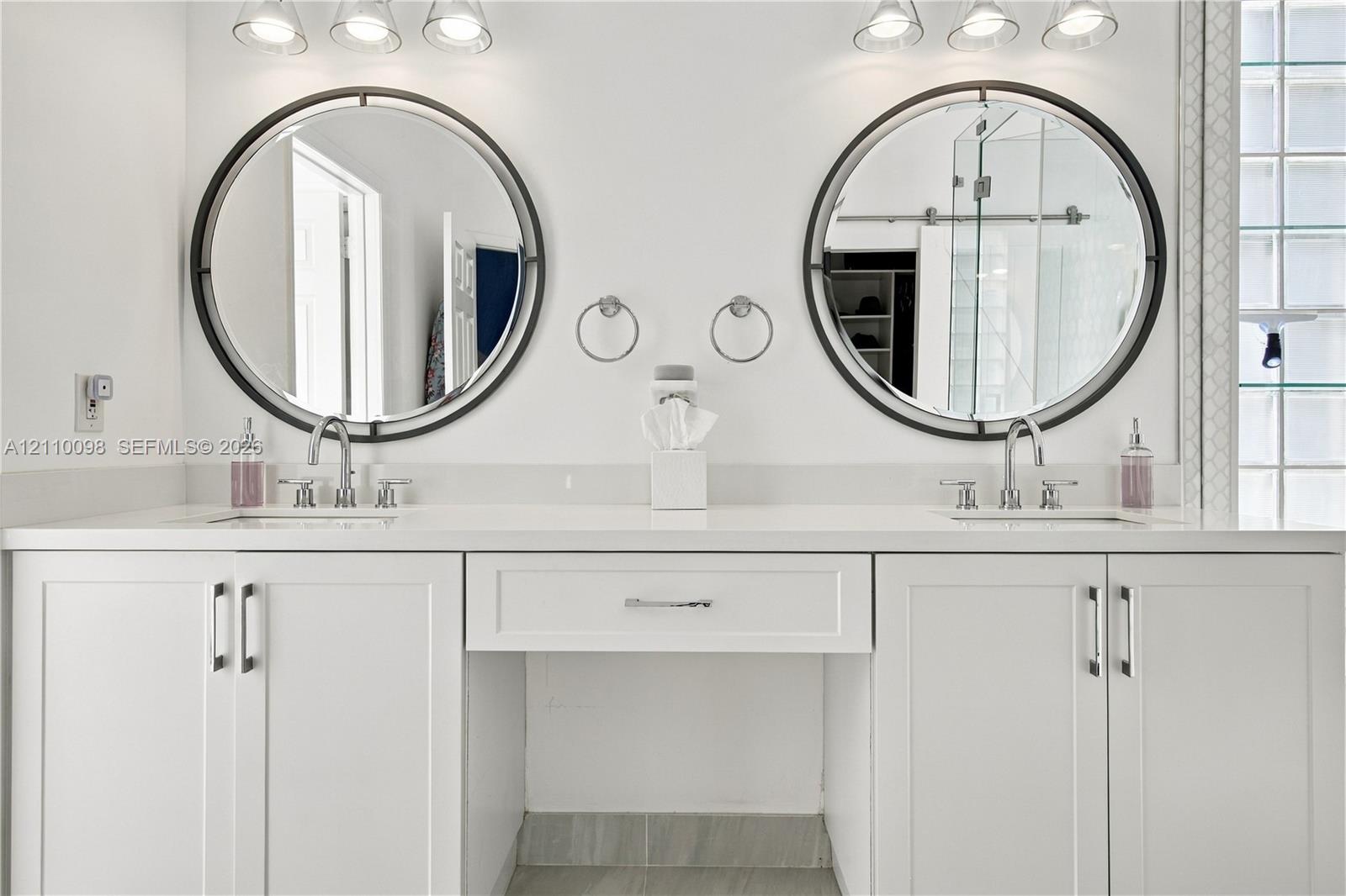 Dual-sink vanity featuring white cabinetry, solid surface countertop, two circular mirrors with dark frames, and chrome fixtures