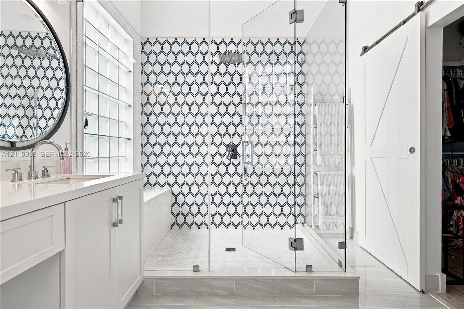 Walk-in shower featuring glass paneling and patterned tile surround, white cabinetry with light-toned countertop, round wall-mounted mirror, and white sliding barn door