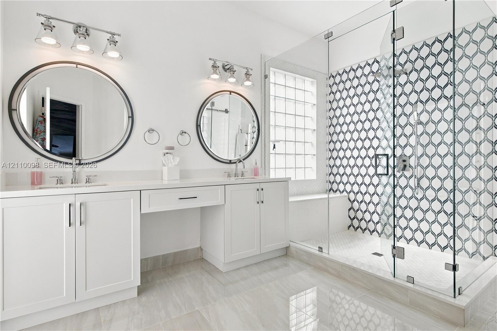 Double vanity featuring white cabinetry, oval mirrors, dual three-light fixtures, and a walk-in glass-enclosed shower with patterned tile surround