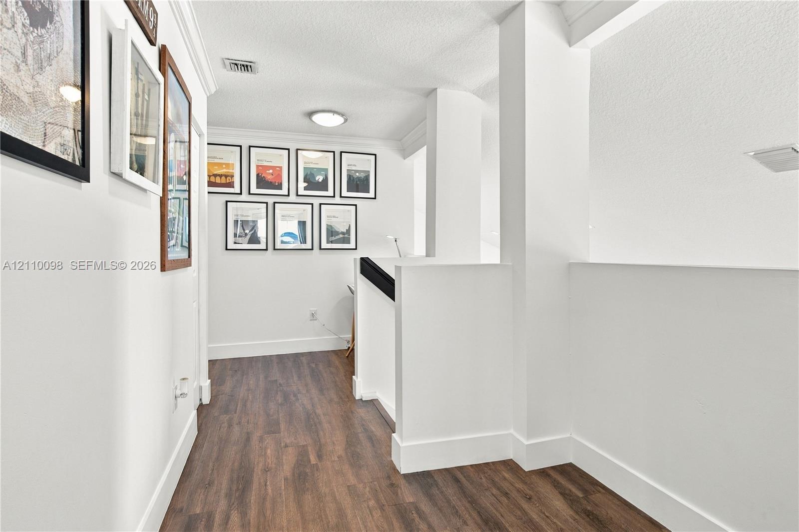 Hallway featuring wood-finish flooring, crisp white walls, crown molding, and a staircase with a solid-surfaced wall and a contrasting handrail