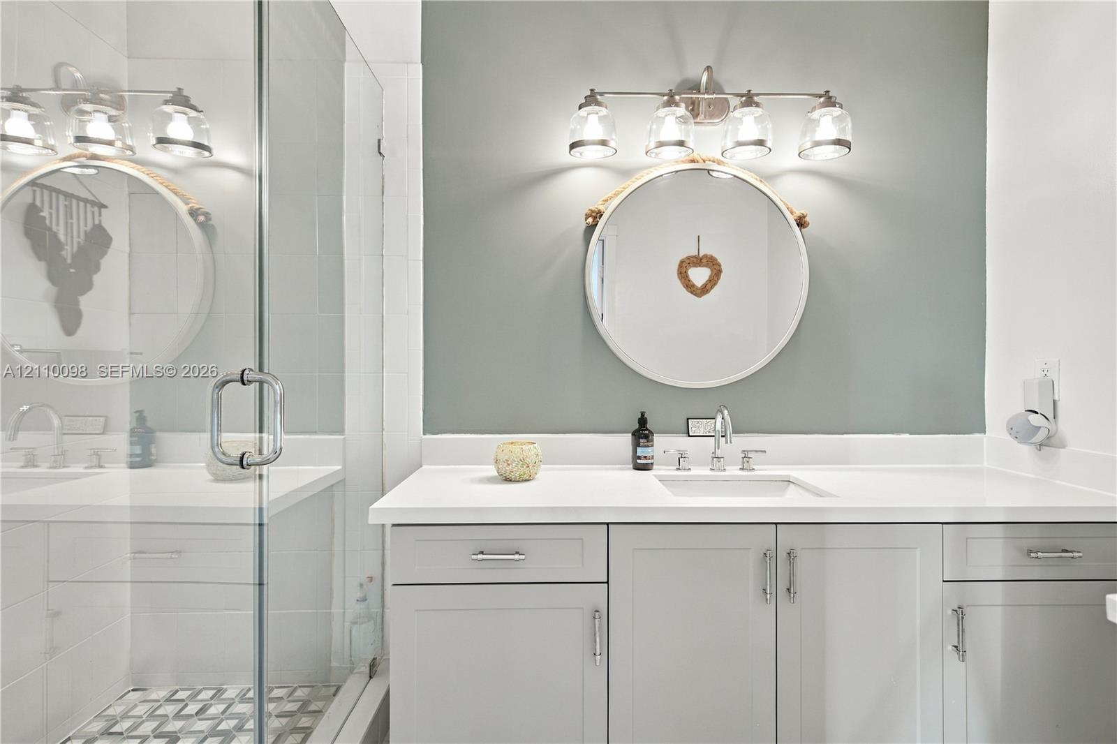 Bathroom featuring a vanity with light gray cabinetry, white solid-surface countertop, rectangular undermount sink, four-light vanity fixture, and a glass-enclosed shower with patterned tile flooring