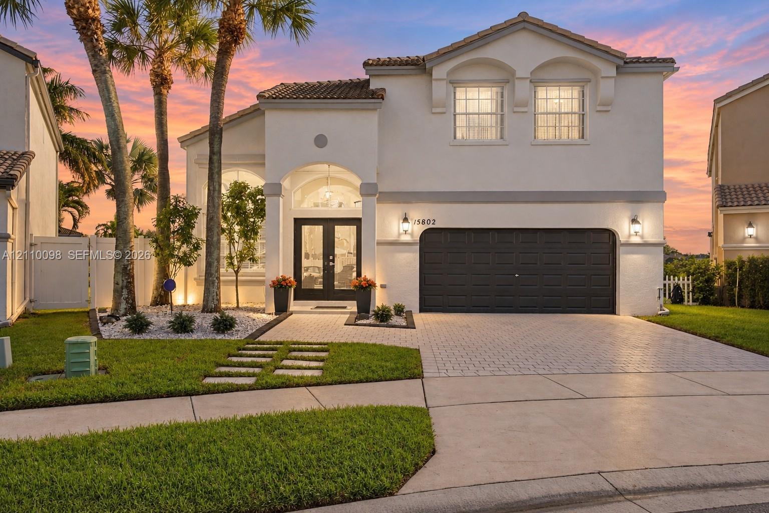 Two-story stucco exterior featuring a paved driveway, arched entryway, double-glass front doors, and decorative landscaping