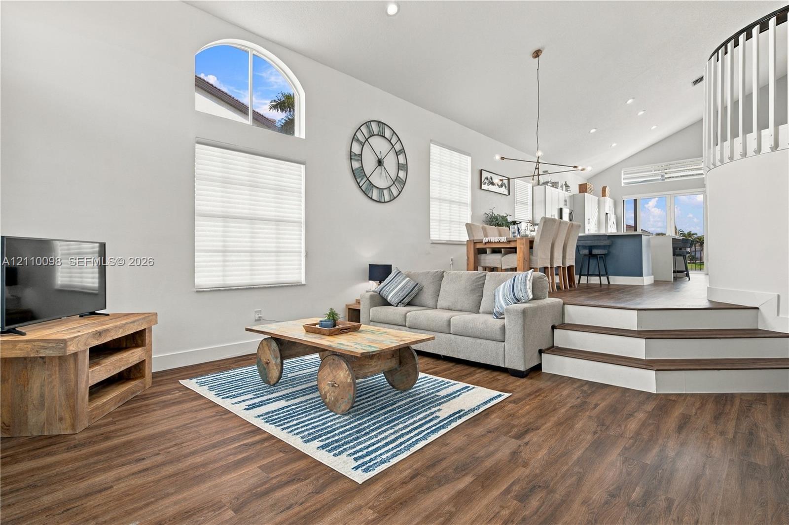 Open-concept living space featuring vaulted ceilings, wood-finish flooring, an arched window, and a multi-level staircase with white balusters