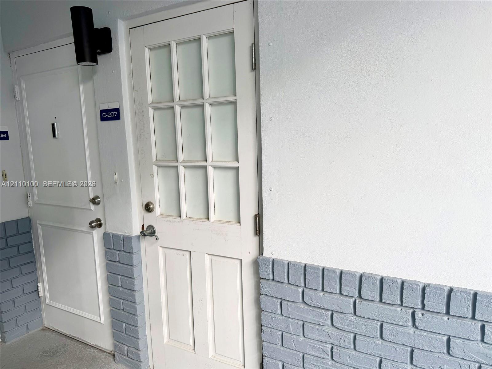 Exterior entry featuring a multi-light glass pane door, paneled solid door, cylindrical wall-mounted sconce, and painted brick wainscoting