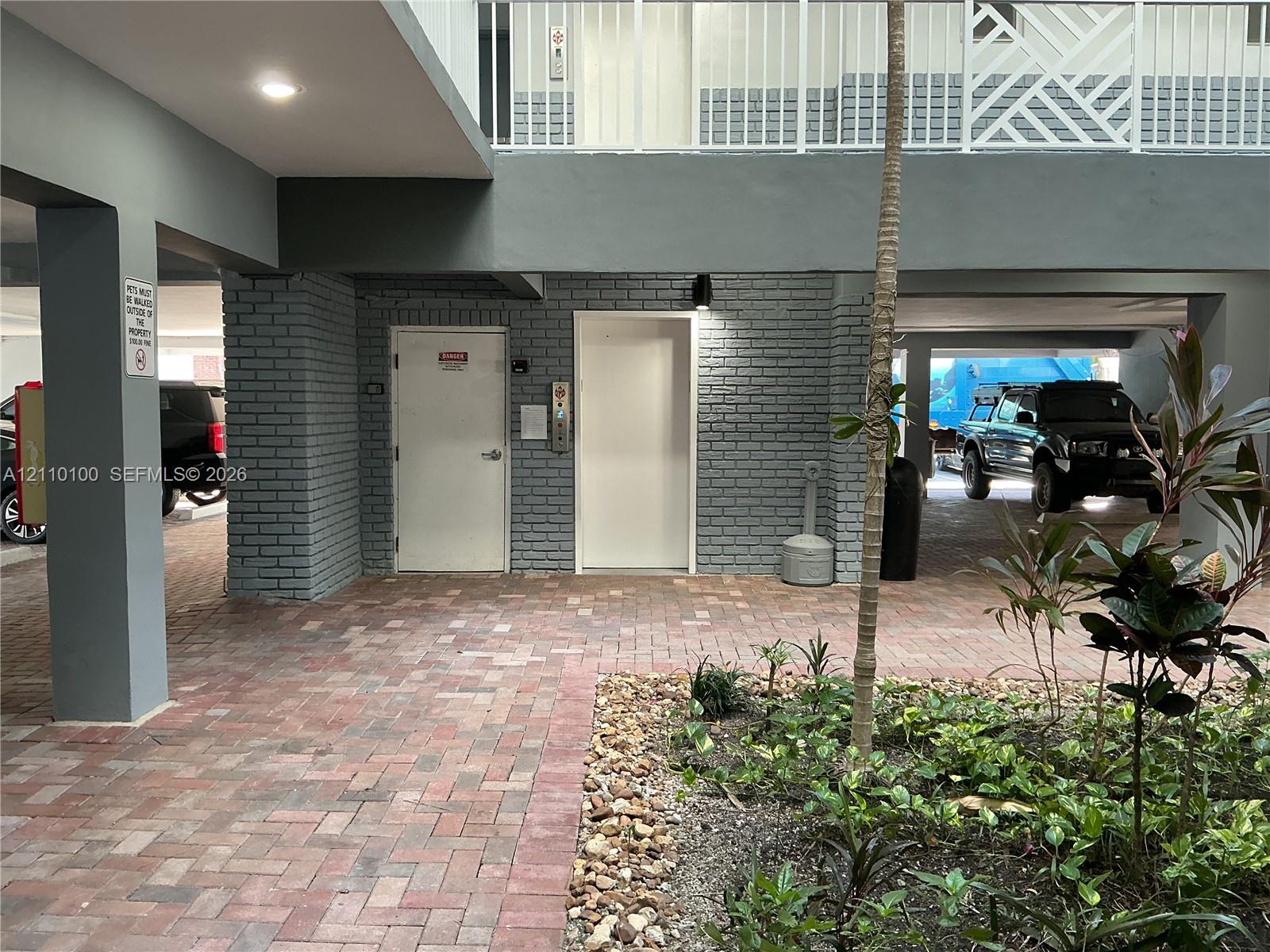 Covered ground-level parking area featuring paver flooring, painted brick structural columns, an elevator, and landscaped garden beds