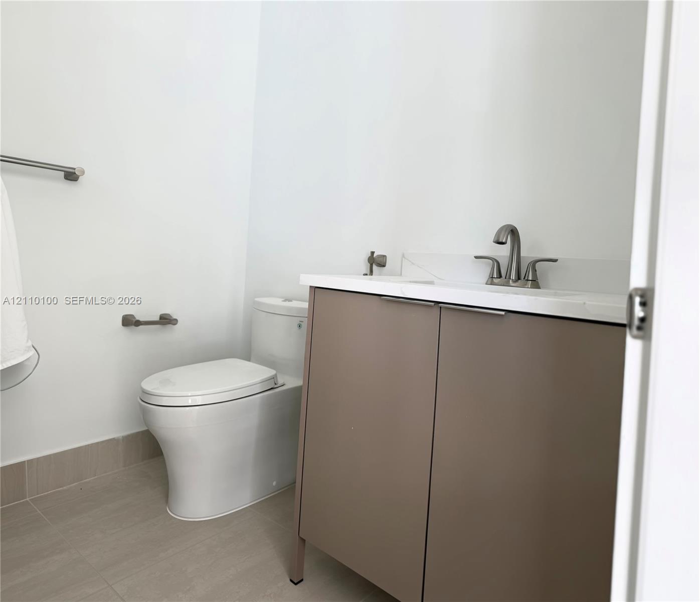 Modern vanity with a neutral-toned cabinet, white stone-composite countertop, dual-handle metal faucet, and floor-mounted commode, complemented by tile flooring