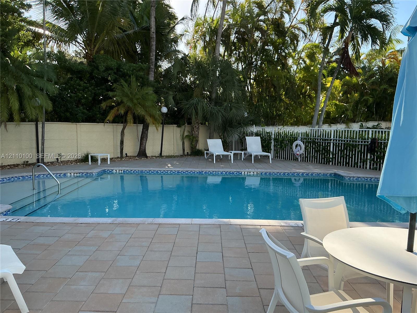 In-ground swimming pool with built-in stairs, surrounded by a paved patio, perimeter fencing, and mature palm trees