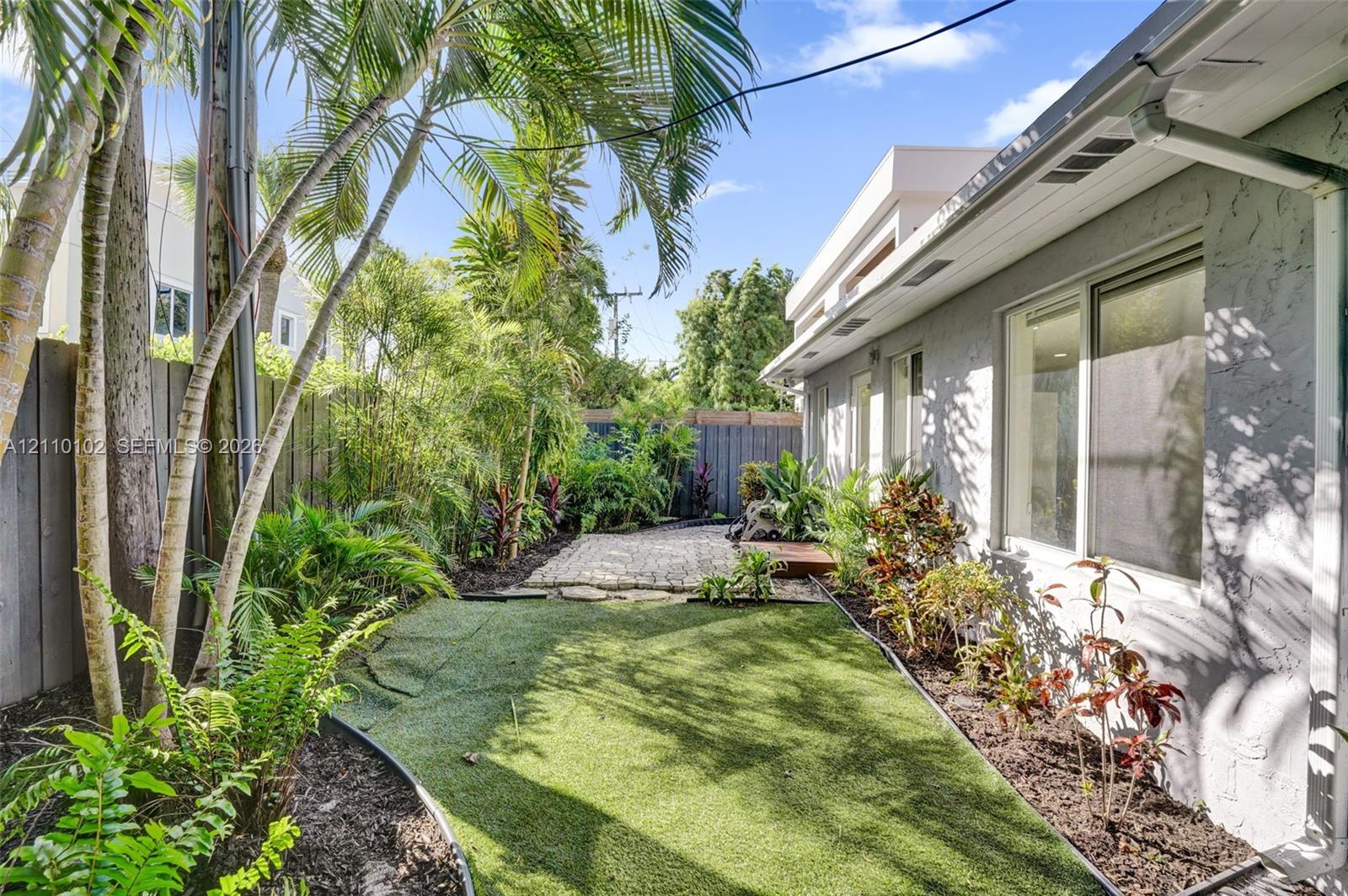 Side yard featuring artificial turf, stone paver accents, established tropical landscaping, and full-height wood fencing
