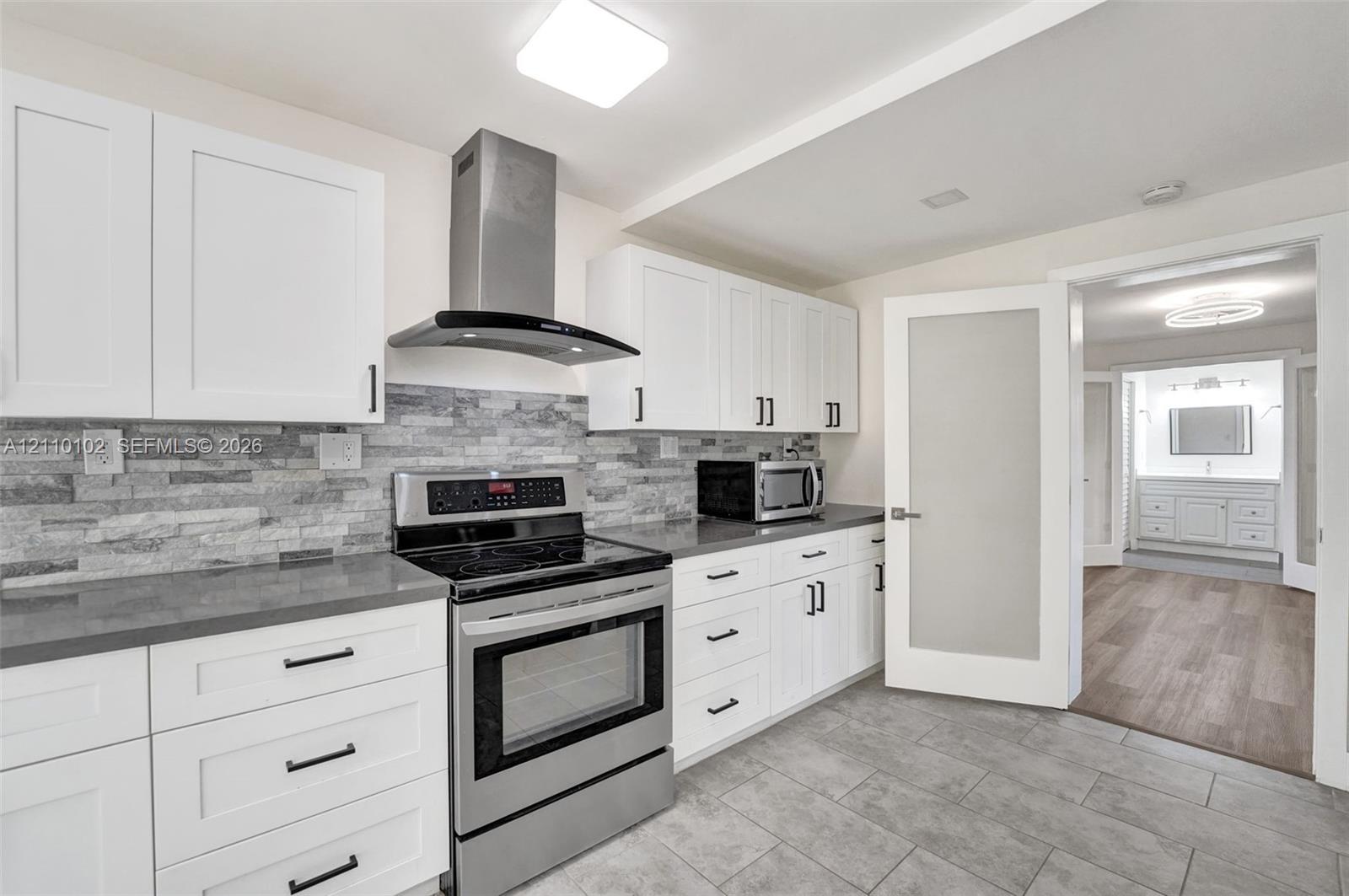 Kitchen featuring white shaker cabinetry, stainless steel appliances including a range and range hood, stone-finish backsplash, solid-surface countertops, and tiled flooring