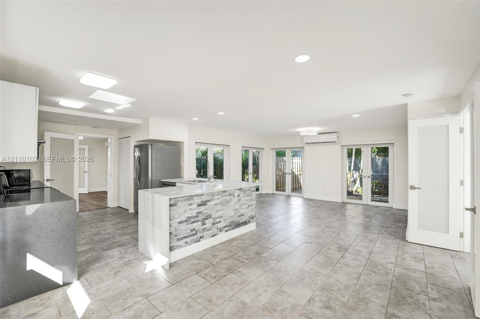 Open-concept kitchen featuring an island with stone-tile cladding and quartz countertops, gray ceramic tile flooring, recessed lighting, and multiple sets of French doors