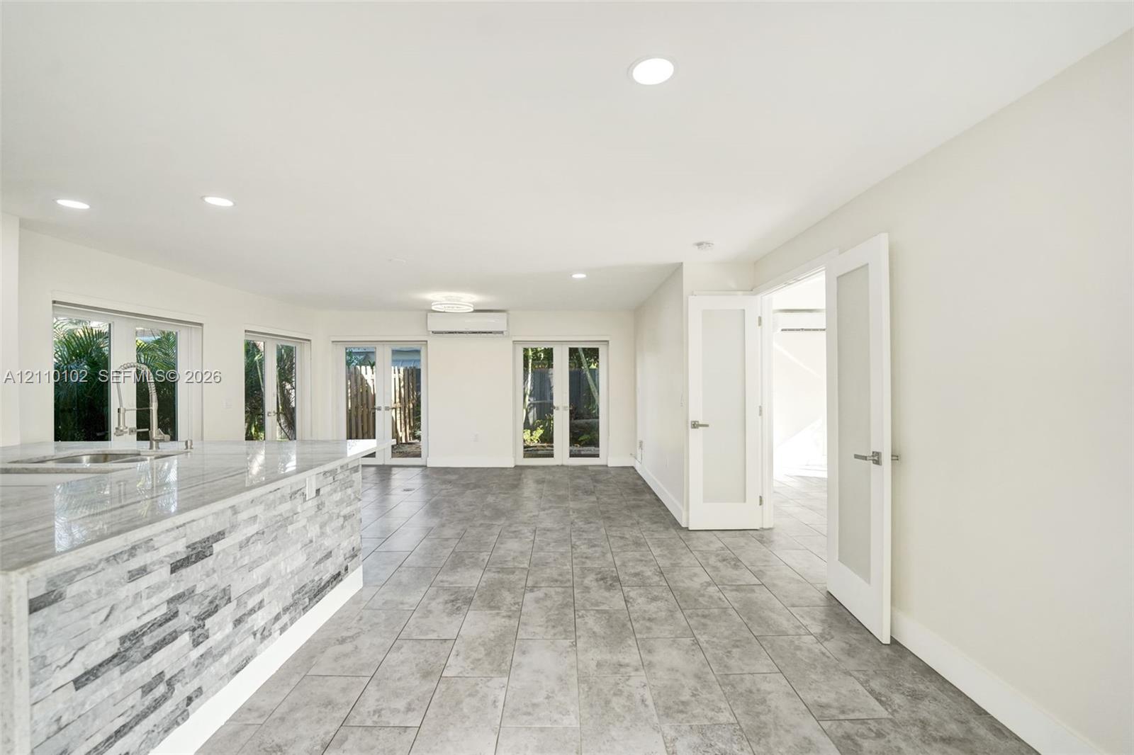 Open-concept interior featuring large-format tile flooring, stone-veneer kitchen island base, recessed lighting, wall-mounted climate control unit, and glass-paneled entry doors