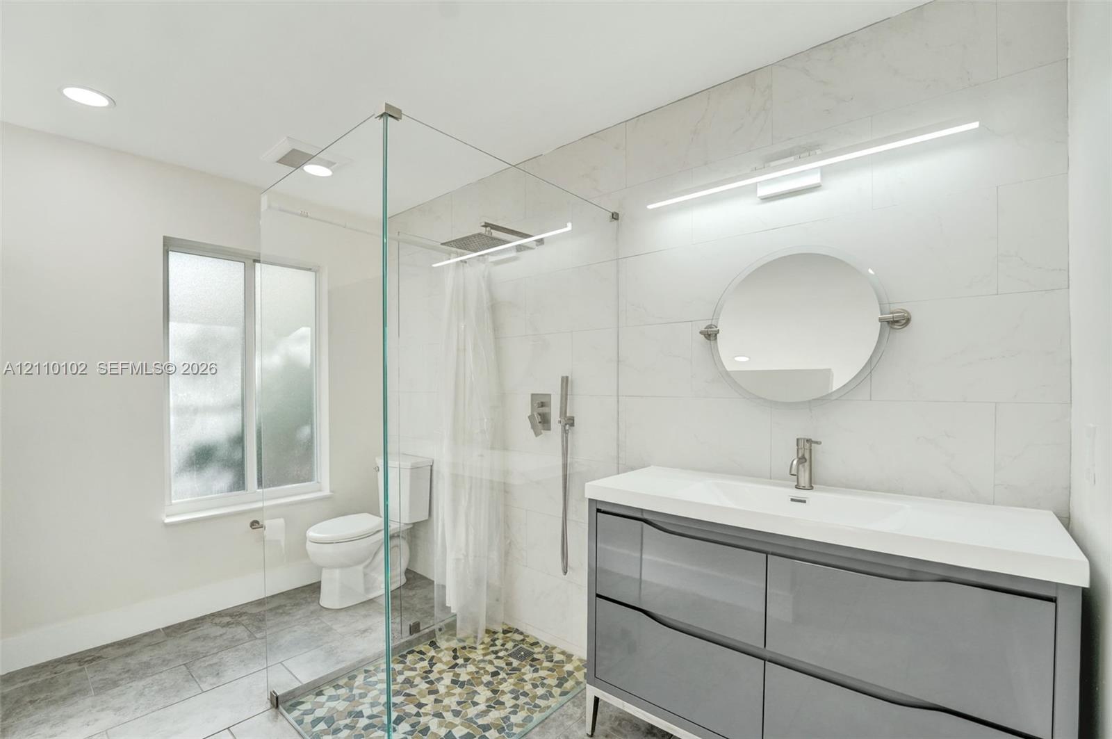 Floating vanity with dual soft-close drawers and integrated countertop, glass-enclosed shower with rainfall showerhead, pebble-tiled shower floor, and large-format wall tiling
