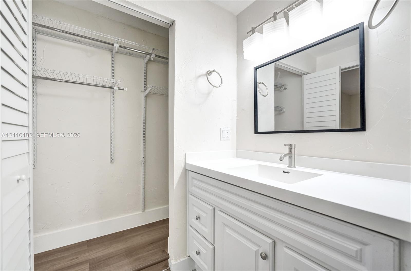 Bathroom vanity featuring a white cabinet, integrated sink, solid-surface countertop, and rectangular mirror with a black frame, accompanied by an open closet with wire shelving and wood-finish flooring