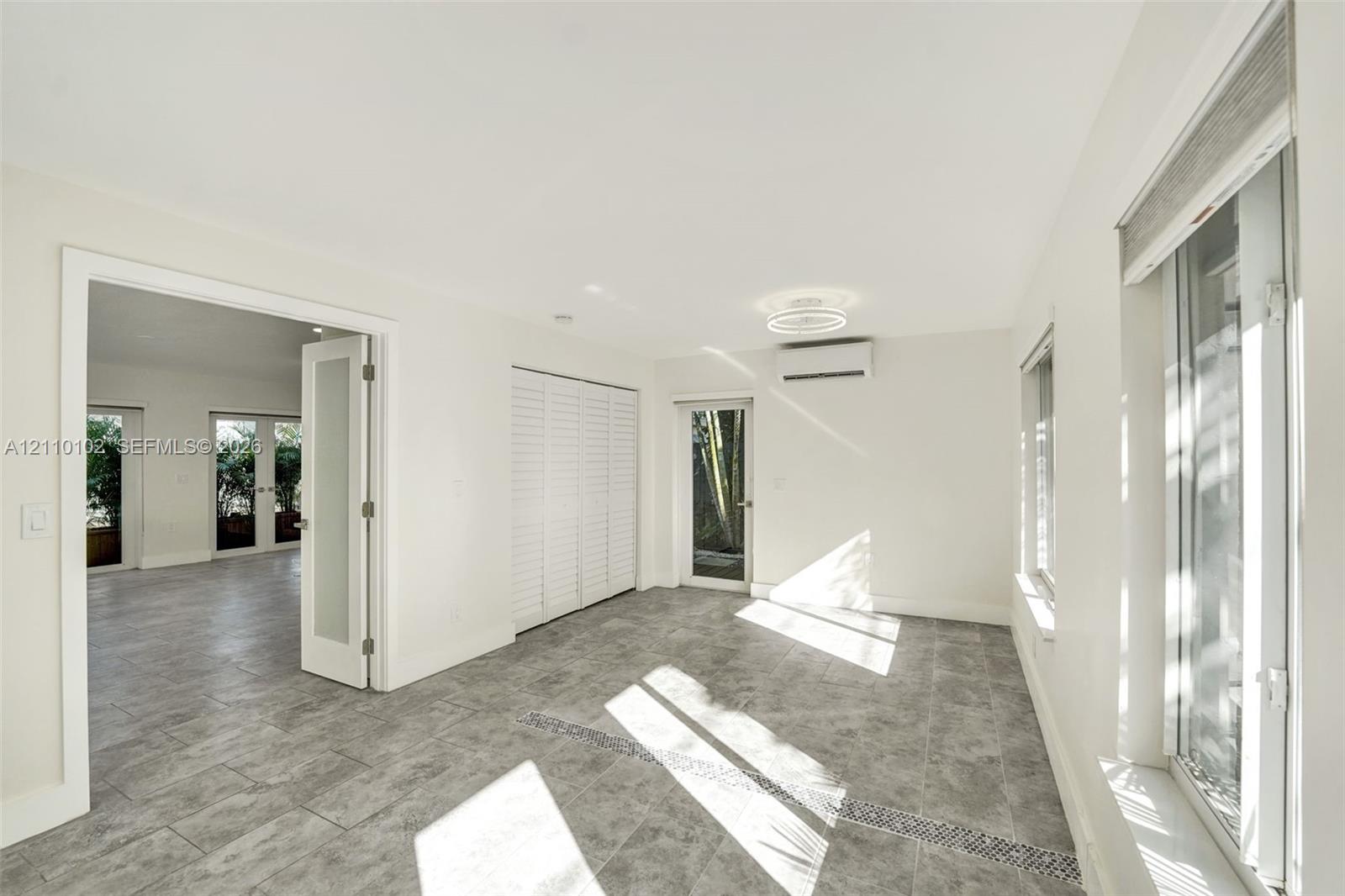 Interior space featuring large-format tile flooring, double glass-paneled doors, built-in closet cabinetry with louvered doors, wall-mounted air conditioning unit, and flush-mount LED circular ceiling light