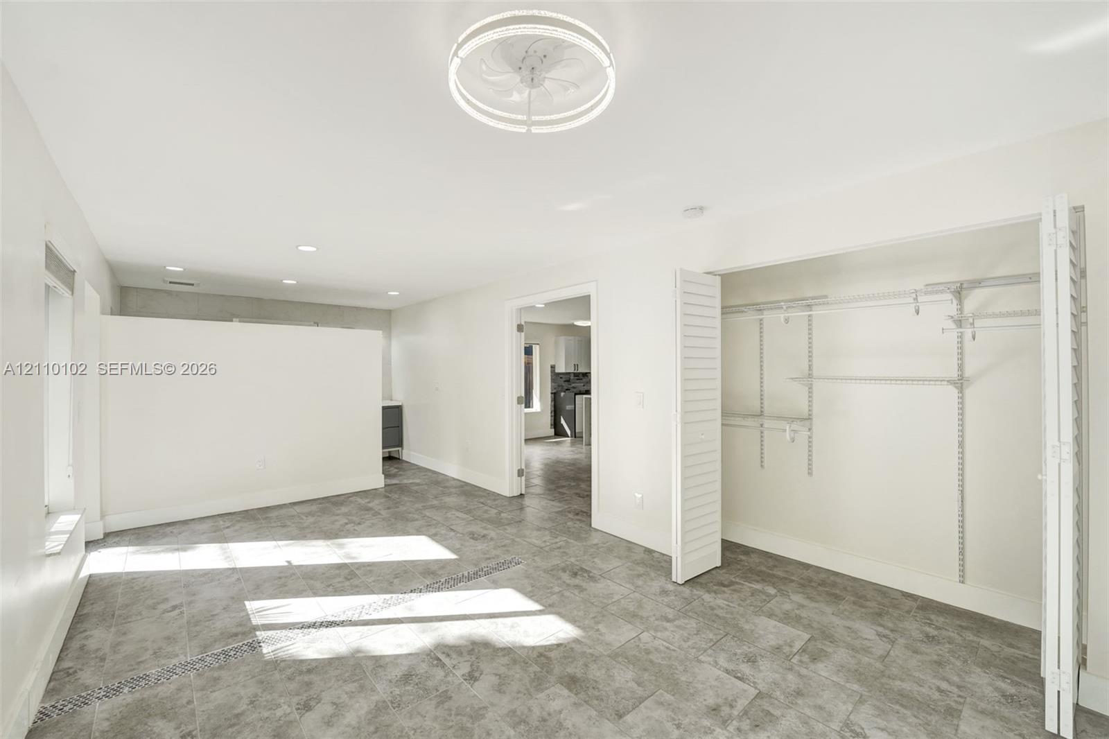 Room featuring large-format tile flooring, a contemporary circular flush-mount ceiling fixture, a partial privacy wall, and a closet with ventilated wire shelving behind louvered doors